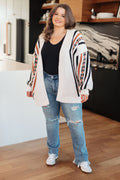 Woman wearing a patterned cardigan, black top, and ripped jeans in a room with wooden flooring.