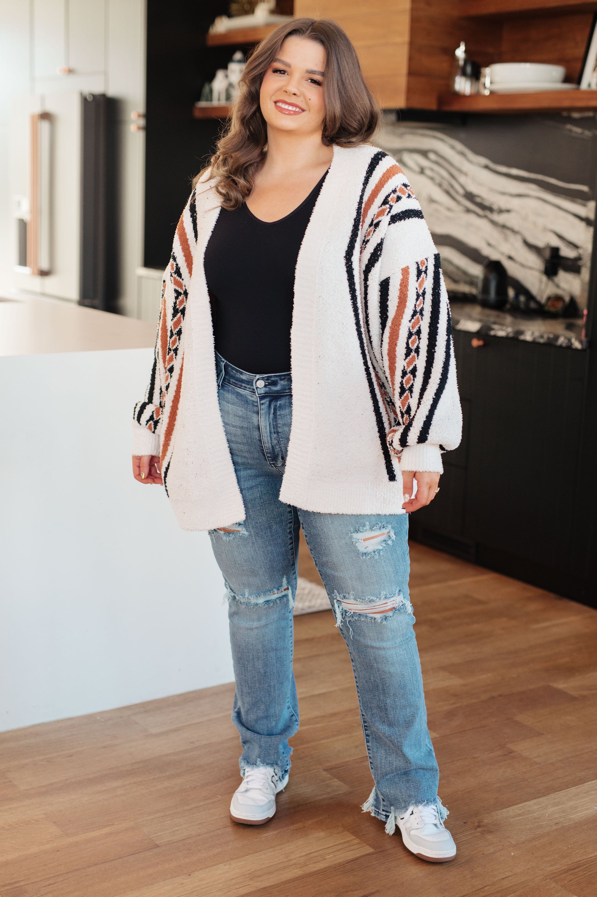 Woman wearing a patterned cardigan, black top, and ripped jeans in a room with wooden flooring.